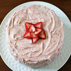 A round cake with pink frosting is topped with sliced strawberries arranged in a star shape, served on a white plate against a wooden background.