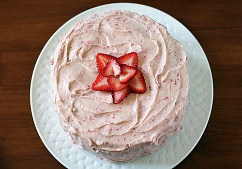 A round cake with pink strawberry frosting is displayed on a white plate, topped with a decorative arrangement of sliced strawberries in the center.