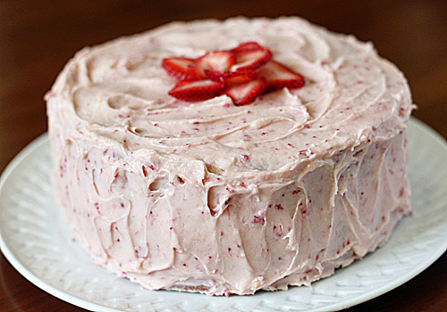 A round cake covered in pink strawberry frosting, topped with sliced fresh strawberries arranged in a flower shape, sits on a white plate.