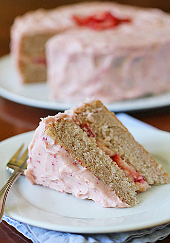 A slice of layered cake with pink frosting and a hint of strawberry filling sits on a white plate with a fork. The rest of the cake is in the background, topped with more pink frosting.