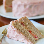 A slice of layered cake with pink frosting and visible strawberry pieces rests on a white plate with a fork, while the remaining cake sits in the background on another plate.