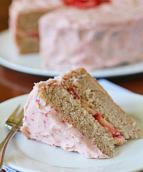 A slice of layered cake with pink frosting and visible strawberry pieces rests on a white plate with a fork, while the remaining cake sits in the background on another plate.