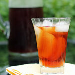 A glass of iced tea with ice cubes sits on a folded yellow and white napkin, with a pitcher of iced tea blurred in the background. The setting appears to be outdoors.