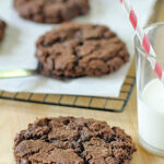 A close-up of a large, chocolate cookie on a wooden surface, with a glass of milk and a red-striped straw beside it. More cookies are cooling on a rack lined with parchment paper in the background.