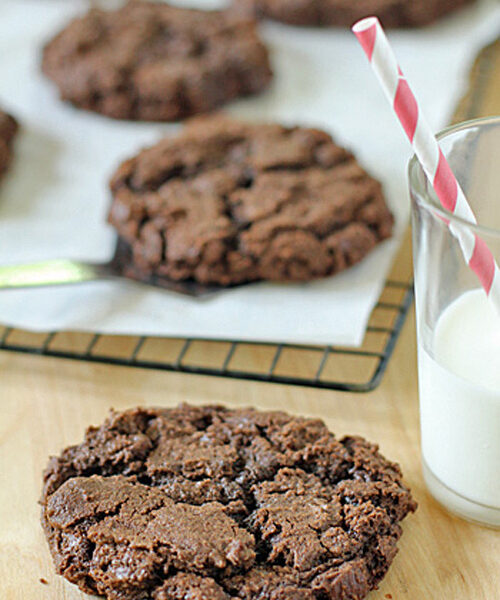 A close-up of a large, chocolate cookie on a wooden surface, with a glass of milk and a red-striped straw beside it. More cookies are cooling on a rack lined with parchment paper in the background.