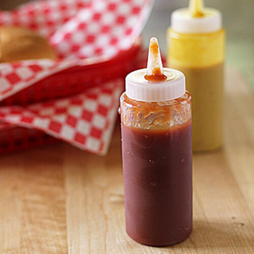A clear squeeze bottle of red ketchup sits on a wooden surface, with a yellow mustard bottle in the background and a red-and-white checkered cloth nearby.