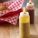 A squeeze bottle of yellow mustard sits on a wooden surface, with a bottle of red sauce and a red-checkered food basket blurred in the background.