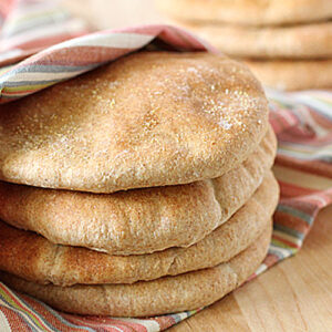 A stack of round, golden-brown pita breads rests partially wrapped in a colorful striped cloth on a wooden surface. More pita breads are visible in the background.