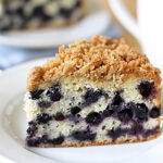 A slice of blueberry crumb cake with streusel topping sits on a white plate. The cake has a light, fluffy texture with visible blueberries throughout. A blurred cup and another cake slice are in the background.