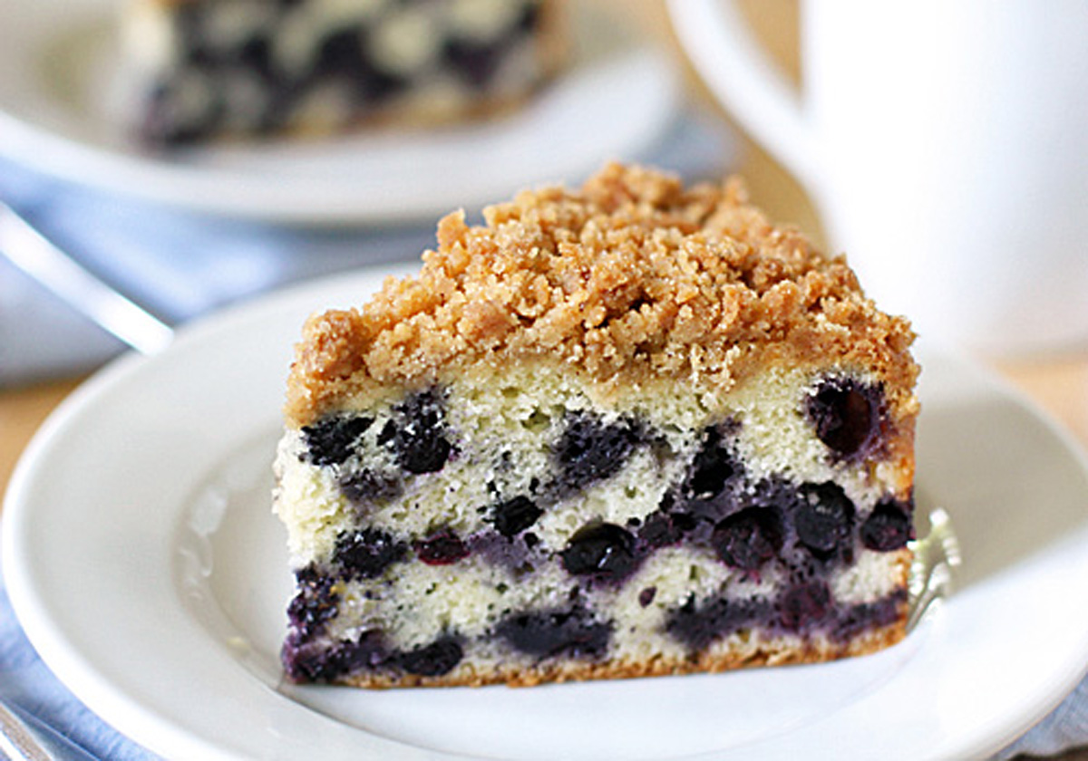 A slice of blueberry crumb cake with streusel topping sits on a white plate. The cake has a light, fluffy texture with visible blueberries throughout. A blurred cup and another cake slice are in the background.