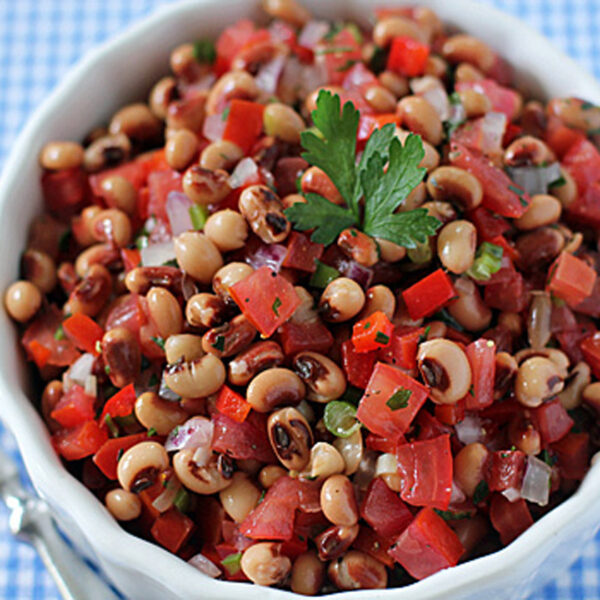 A white bowl filled with black-eyed pea salad mixed with diced tomatoes, red bell peppers, onions, and garnished with parsley, placed on a blue and white checkered tablecloth.