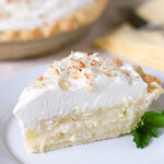 A slice of coconut cream pie topped with whipped cream and toasted coconut flakes sits on a white plate, garnished with a sprig of mint. A pie dish and utensils are visible in the background.