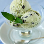 Two scoops of mint chocolate chip ice cream in a glass dessert dish, garnished with a fresh mint leaf, served on a white saucer with a silver spoon beside it.