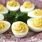 A plate of deviled eggs, each half filled with a creamy, yellow yolk mixture, displayed on a decorative maroon and white dish with a sprig of fresh herbs in the center.