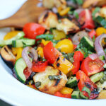 A close-up of a colorful panzanella salad with grilled bread, cherry tomatoes, cucumbers, red onions, yellow bell peppers, capers, and fresh basil in a white bowl, with a wooden serving fork in the background.