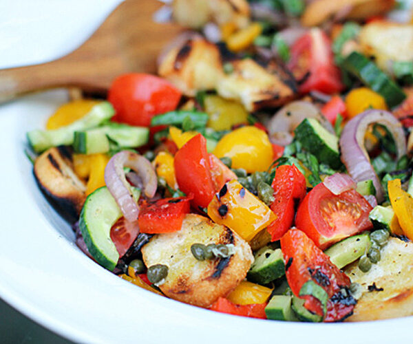 A close-up of a colorful panzanella salad with grilled bread, cherry tomatoes, cucumbers, red onions, yellow bell peppers, capers, and fresh basil in a white bowl, with a wooden serving fork in the background.