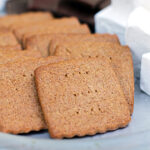 Square graham crackers are arranged on a plate next to a stack of fluffy white marshmallows and pieces of chocolate, ready for making smores.