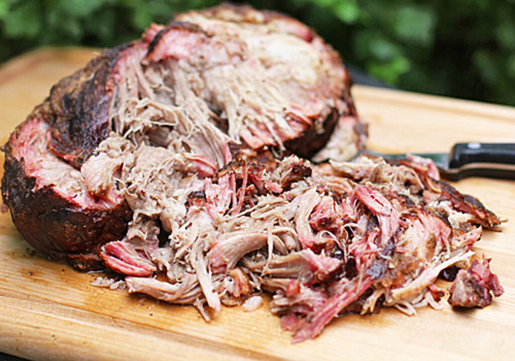 A large portion of smoked, shredded pulled pork sits on a wooden cutting board, with a knife nearby. The meat looks juicy and tender with a mix of crispy, browned edges and pink interior.