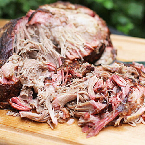 A large portion of smoked, shredded pulled pork sits on a wooden cutting board, with a knife nearby. The meat looks juicy and tender with a mix of crispy, browned edges and pink interior.