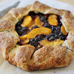 A rustic fruit galette with a golden-brown, flaky crust is filled with sliced peaches and blueberries, sitting on parchment paper next to a knife.