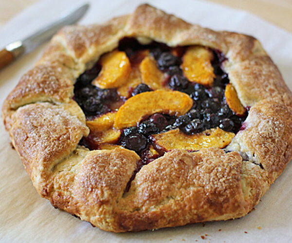 A rustic fruit galette with a golden-brown, flaky crust is filled with sliced peaches and blueberries, sitting on parchment paper next to a knife.