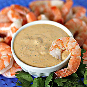 A bowl of creamy dipping sauce is surrounded by cooked shrimp, arranged in a circle on a blue plate. Fresh cilantro leaves are placed near the bowl for garnish.