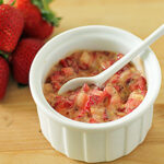 A white ramekin filled with a creamy strawberry mixture, accompanied by a white spoon. Fresh whole strawberries are placed beside the ramekin on a wooden surface.