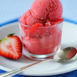 A glass cup filled with bright pink strawberry sorbet sits on a white plate, accompanied by a halved strawberry and a silver spoon on a blue napkin.
