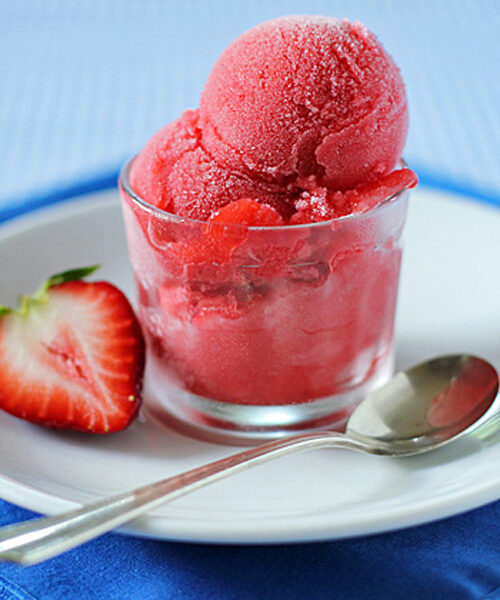A glass cup filled with bright pink strawberry sorbet sits on a white plate, accompanied by a halved strawberry and a silver spoon on a blue napkin.