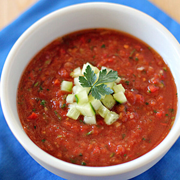 A white bowl filled with gazpacho soup, topped with diced cucumber and a sprig of parsley, sits on a blue cloth next to a spoon with a wooden handle.