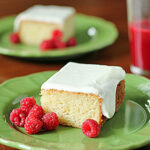 A square piece of frosted cake on a green plate with fresh raspberries, and a glass of red juice in the background. Another plate with cake and raspberries is visible further back.