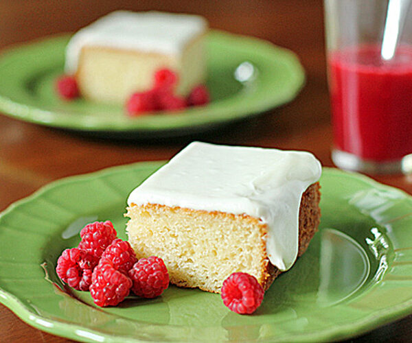 A square piece of frosted cake on a green plate with fresh raspberries, and a glass of red juice in the background. Another plate with cake and raspberries is visible further back.
