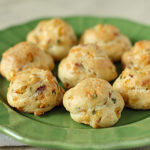 A green plate holds eight round, golden-brown savory pastry puffs with visible bits of herbs and cheese baked into them. The background is softly blurred.