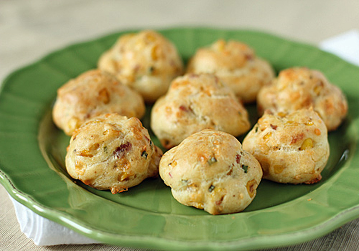 A green plate holds eight round, golden-brown savory pastry puffs with visible bits of herbs and cheese baked into them. The background is softly blurred.