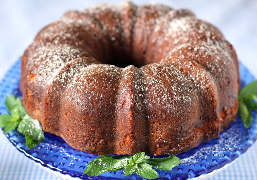 A golden-brown bundt cake dusted with powdered sugar sits on a blue plate, garnished with fresh mint leaves around the base.