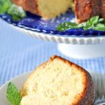 A slice of golden brown bundt cake sits on a white plate with a mint garnish, in front of the remaining cake on a blue patterned cake stand. A fork rests beside the slice.