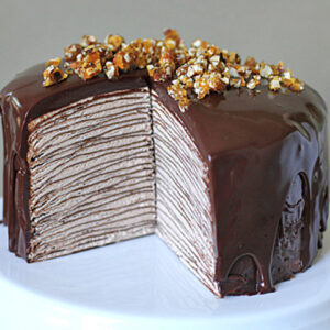 A tall chocolate crepe cake with many thin layers, covered in glossy chocolate ganache and topped with crushed praline, sits on a white cake stand with a large slice removed to show the layered interior.
