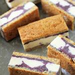 Rectangular ice cream sandwiches with a layer of blueberry swirl ice cream between two pieces of golden brown cake, arranged on a gray surface.