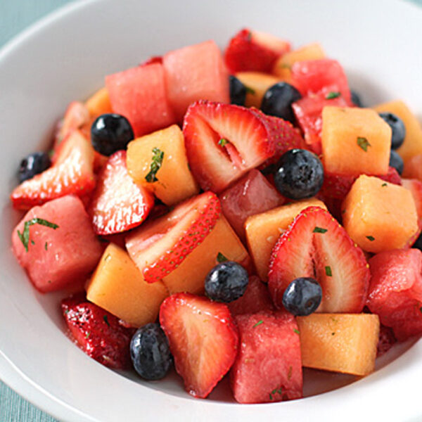 A white bowl filled with a colorful fruit salad, including sliced strawberries, blueberries, cubed cantaloupe, and cubed watermelon, sits on a table next to a spoon.