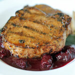 A grilled pork chop with seared grill marks sits atop a bed of dark red berry sauce on a white plate, accompanied by a fork.