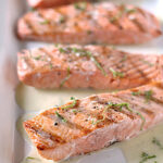 Four pieces of grilled salmon fillet on a white plate, garnished with herbs and served with a light creamy sauce. The salmon appears tender and freshly cooked.