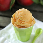 A scoop of peach sorbet sits in a light green cup on a white napkin, with a matching green spoon beside it. Out-of-focus peaches and green leaves are visible in the background.