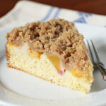 A slice of crumb-topped peach coffee cake on a white plate with a fork, set on a light-colored cloth with blue patterns.