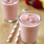 Two glasses of creamy pink smoothie sit on a wooden surface, with two red-and-white striped straws beside them. In the background, a banana and two strawberries are slightly out of focus.