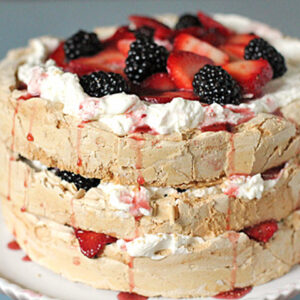 A two-layer pavlova cake topped with whipped cream, sliced strawberries, and blackberries, with berry sauce dripping down the sides, displayed on a white plate.