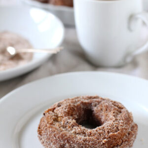 A cinnamon sugar-coated donut on a white plate with a cup of coffee and a dish of cinnamon sugar in the background.