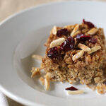 A square serving of baked oatmeal sits on a white plate, topped with slivered almonds and dried cranberries. A fork and napkin are placed to the left of the plate.