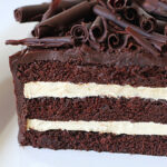 A close-up of a layered chocolate cake with two layers of light cream filling, topped with chocolate curls and a glossy chocolate glaze.