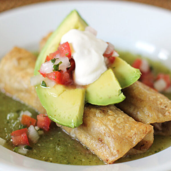 Three crispy rolled tacos on a plate with green salsa, topped with avocado slices, pico de gallo, and a dollop of sour cream.