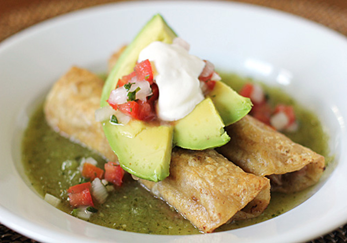 Three crispy rolled tacos on a plate with green salsa, topped with avocado slices, pico de gallo, and a dollop of sour cream.